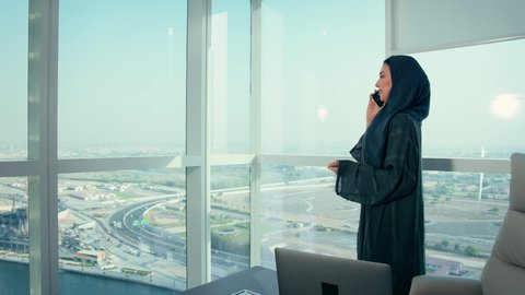 Making a phone call at the company headquarters, auditing and analyzing project data, following up on the chart plan, an Emirati Gulf Arab woman analyzing data on a laptop, economic growth and financial investment, a Saudi woman wearing a hijab and abaya, background of skyscrapers and towers.