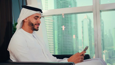 تبادل الأحاديث والأخبار عبر الهاتف، شركات الامارات العربية المتحدة، تصوير مقرب لايدي رجل عربي خليجي اماراتي يرتدي الكندورة والغترة البيضاء يحمل هاتف محمول، إنجاز مهام العمل عن بعد، إدارة شؤون الأعمال وتنظيمها عبر اللابتوب، خلفية ابراج وناطحات سحاب