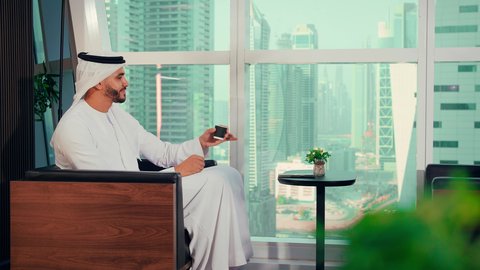 احتساء المشروب المفضل،  قضاء وقت ممتع و هادئ، مفهوم الطاقة والإيجابية، الحيوية والنشاط عند شرب الكافيين، رجل عربي خليجي اماراتي يرتدي الكندورة و الغتره يجلس على كرسي بجانب النافذة يحمل بيده فنجان من القهوه، الاستمتاع بتناول المشروبات الساخن