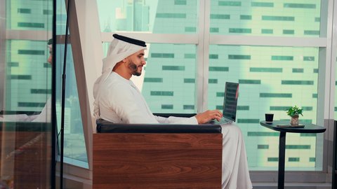 دمج العمل مع التقنية والتكنولوجيا، تصوير مقرب لرجل عربي خليجي اماراتي يرتدي الكندورة والغترة البيضاء يستخدم الحاسوب المحمول، إنجاز مهام العمل عن بعد، إدارة شؤون الأعمال وتنظيمها عبر اللابتوب، ايماءات السعادة والسرور، مواقع التواصل الإجتماعي، خلفية ابراج وناطحات سحاب