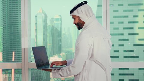 التأمل بمعالم المدينة، تصوير مقرب لرجل عربي خليجي اماراتي يرتدي الكندورة والغترة البيضاء يستخدم الحاسوب المحمول، إنجاز مهام العمل عن بعد، إدارة شؤون الأعمال وتنظيمها عبر اللابتوب، ايماءات السعادة والسرور، مواقع التواصل الإجتماعي، خلفية ابراج وناطحات سحاب