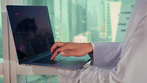 الايدي السريعة على لوحة المفاتيح، تصوير مقرب لرجل عربي خليجي اماراتي يرتدي الكندورة والغترة البيضاء يستخدم الحاسوب المحمول، إنجاز مهام العمل عن بعد، إدارة شؤون الأعمال وتنظيمها عبر اللابتوب، ايماءات السعادة والسرور، مواقع التواصل الإجتماعي، خلفية ابراج وناطحات سحاب