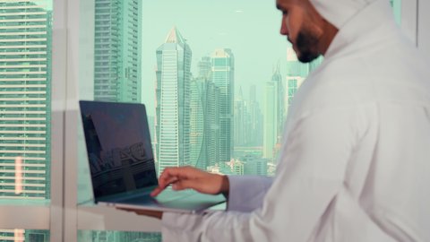 The use of a modern and advanced technical device, a close-up shot of an Arab Gulf Emirati man wearing a kandura and a white ghutrah using a laptop, accomplishing remote work tasks, managing and organizing business affairs through the laptop, gestures of happiness and joy, social media, with a background of towers and skyscrapers.
