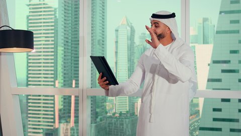 رفع اليد بتعابير القاء التحية والسلام، شركات الامارات العربية المتحدة، تصوير مقرب لايدي رجل عربي خليجي اماراتي يرتدي الكندورة والغترة البيضاء يحمل جهاز لوحي، إنجاز مهام العمل عن بعد، إدارة شؤون الأعمال وتنظيمها عبر اللابتوب، خلفية ابراج وناطحات سحاب