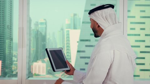 The concept of remote project management, companies in the United Arab Emirates, a close-up shot of the hands of an Emirati Arab man wearing a kandura and a white ghutrah holding a tablet, accomplishing work tasks remotely, managing and organizing business affairs via a laptop, with a background of towers and skyscrapers.