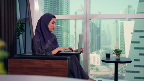 أداء المهام والاعمال المكتبيه داخل مقر الشركه، متابعة مخطط الرسم البياني، تصوير مقرب لامراه عربيه خليجيه اماراتيه تقوم بالعمل على الحاسب المحمول، النمو الاقتصادي والاستثمار المالي، سيدة سعودية ترتدي الحجاب والعباءة، خلفية ناطحات سحاب وابراج