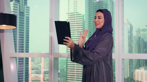القاء التحية عبرمكالمة فيديو ، متابعة مخطط الرسم البياني، تصوير مقرب لامراه عربيه خليجيه اماراتيه تقوم بالعمل على الجهاز اللوحي ، النمو الاقتصادي والاستثمار المالي، سيدة سعودية ترتدي الحجاب والعباءة، خلفية ناطحات سحاب وابراج، الاناقة ولاهتمام بالمظهر الخارجي