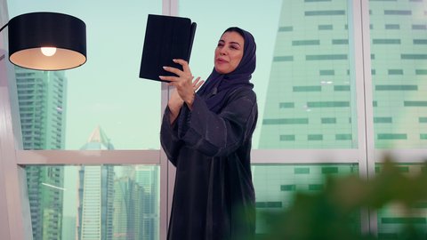 تواصل عبرمكالمة فيديو باستخدام التابلت، متابعة مخطط الرسم البياني، تصوير مقرب لامراه عربيه خليجيه اماراتيه تقوم بالعمل على الجهاز اللوحي ، النمو الاقتصادي والاستثمار المالي، سيدة سعودية ترتدي الحجاب والعباءة، خلفية ناطحات سحاب وابراج، الاناقة ولاهتمام بالمظهر الخارجي