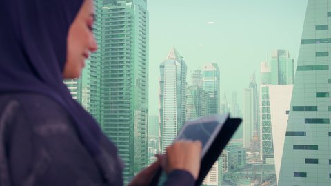 انهاء العمل عن بعد، متابعة مخطط الرسم البياني، تصوير مقرب لامراه عربيه خليجيه اماراتيه تقوم بالعمل على الجهاز اللوحي ، النمو الاقتصادي والاستثمار المالي، سيدة سعودية ترتدي الحجاب والعباءة، خلفية ناطحات سحاب وابراج، الاناقة ولاهتمام بالمظهر الخارجي