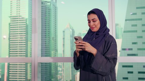 تصفح مواقع التواصل الاجتماعي، متابعة مخطط الرسم البياني، تصوير مقرب لامراه عربيه خليجيه اماراتيه تقوم بالعمل على الهاتف المحمول، النمو الاقتصادي والاستثمار المالي، سيدة سعودية ترتدي الحجاب والعباءة، خلفية ناطحات سحاب وابراج، الاناقة ولاهتمام بالمظهر الخارجي
