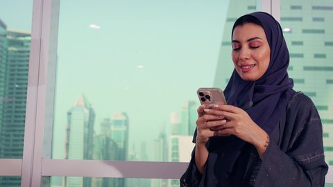 Working on the phone at the company headquarters, browsing social media, following the chart plan, close-up shot of an Arab Gulf Emirati woman, economic growth and financial investment, a Saudi woman wearing a hijab and abaya, background of skyscrapers and towers, elegance and attention to external appearance.