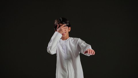 التفاعل مع البيئة المحيطة، بورتريه لولد عربي خليجي سعودي يرتدي الثوب الأبيض يمد يده إلى الأمام ويحجب عينيه بيده الأخرى، تعابير وجه تدل على الحذر والترقب، خلفية سوداء