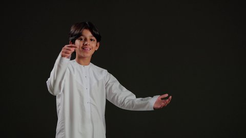 البشاشة والسرور وتحديد موقع معين، حركات تدل على هذا المكان (هنا)، بورتريه لولد عربي خليجي سعودي يرتدي الثوب الابيض يشير بأصابعه نحو الجانب، خلفية سوداء