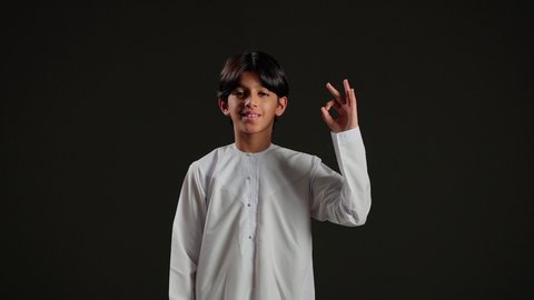 البشاشة ونظرات الحماس والشعور بالرضا, الموافقة والقبول والنصيحة، التعبير عن الجودة والاتقان،بورتريه لولد عربي خليجي سعودي يرتدي الثوب الابيض يرفع يديه بإيماءات الجودة والامتياز والتميز، خلفية سوداء