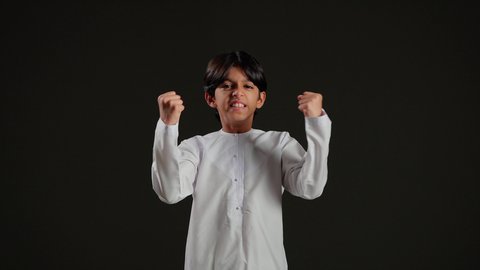 النظر الى الكاميرا بتعابير الفوز والكسب، ايماءات الفرح للانتهاء من انجاز شيء معين، مفهوم النجاح و تحقيق الاهداف، بورتريه لولد عربي خليجي سعودي يرتدي الثوب الابيض يرفع يدية للأعلى بحركات التشجيع والحماس، خلفية سوداء