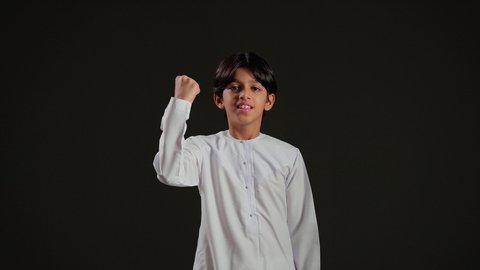 رفع قبضة اليد نحو الأعلى بإيماءات القوة والتحدي، التشجيع بتعابير الحماس، الفرح بالانتصار والفوز،  بورتريه لولد عربي خليجي سعودي يرتدي الثوب الابيض يرفع يده في الهواء بسعادة وسرور، خلفية سوداء