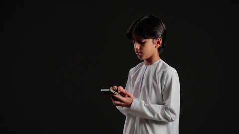 Chatting with family and friends via mobile, browsing social media, a side portrait of a young Arab Gulf Saudi boy wearing a white thobe holding a mobile phone, using a modern and advanced technological device, black background.