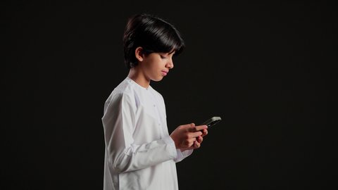 تصفح مواقع التواصل الاجتماعي، الدردشة مع الاهل والاصدقاء عبر الجوال، بورتريه من الجانب لولد عربي خليجي سعودي يرتدي الثوب الابيض  يمسك الهاتف المحمول, استخدام جهاز تقني حديث ومتطور، خلفية سوداء