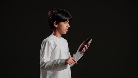 تبادل الأخبار مع الأهل والأصدقاء، إجراء مكالمة هاتفية باستخدام الموبايل، بورتريه لولد عربي خليجي سعودي يرتدي الثوب الابيض يقوم بالتواصل مع الأقارب، تعابير الانفعال و الغضب، خلفية سوداء