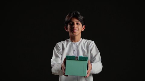 The concept of gratitude and appreciation, giving gifts during holidays and special occasions, expressing love, respect, and admiration, a portrait of an Arab Gulf Saudi boy wearing a white thobe holding a gift box in his hands, black background.