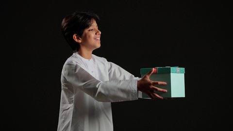 تقديم الهدايا بالاعياد والمناسبات الخاصة، مفهوم الشكر والامتنان، التعبير عن الحب والتقدير والاحترام، بورتريه لولد عربي خليجي سعودي يرتدي الثوب الابيض يحمل بيديه صندوق هدية، خلفية سوداء