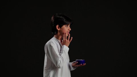 استخدام كريمات ومكونات تجميلية ، بورتريه من الجانب لولد عربي خليجي سعودي يرتدي الثوب التقليدي يمسك بيده كريم المرطب، مفهوم العناية والإهتمام بنضارة وتحسين البشرة، ترطيب وتنظيف وتفتيح الجلد، خلفية سوداء