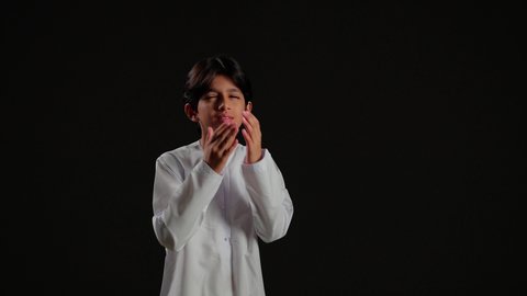 Gestures and facial and hand expressions indicating fear and dread and screaming, a portrait of an Arab Gulf Saudi boy wearing a white thobe with his hands on his face, black background.