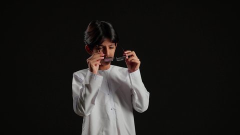 الرؤية من خلال النظارة الطبية، بورتريه لولد عربي خليجي سعودي يرتدي الثوب السعودي يحمل بيده نظارة طبية خاصة بضعف النظر، مفهوم ضعف البصر، قصر وطول النظر، خلفية سوداء