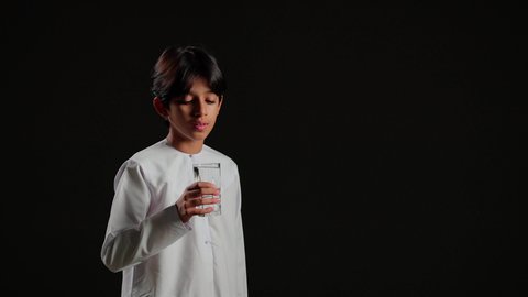 Maintaining the health of the body's immune system, consistently drinking water, feeling thirsty and dehydrated, a portrait of an Arab Gulf Saudi boy wearing a white thobe holding a cup of water, black background.