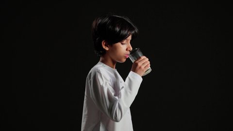 مفهوم العناية والاهتمام بالصحة، المحافظة على سلامة الجهاز المناعي، فوائد مشتقات اللبن المتعددة للجسم والعظام، اتباع نظام غذائي صحي, بورتريه مقرب من الجانب لولد عربي خليجي سعودي يرتدي الثوب الابيض يقوم بشرب كأس من الحليب, خلفية سوداء
