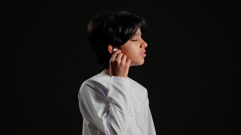 The concept of using mobile device accessories, a modern Bluetooth headset, a close-up portrait of an Emirati Gulf Arab boy wearing a kandura holding his wireless headset in his hands, mobile devices and technology, black background.