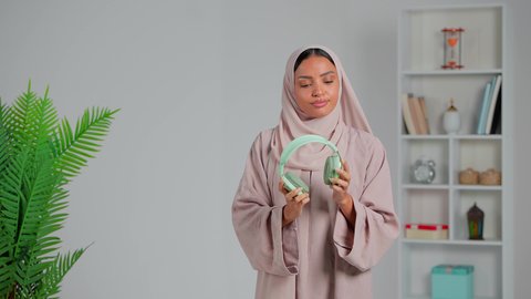 التطور التقني والتكنولوجي, استخدام تقنية الهيدفون، التواصل مع الأهل والأصدقاء، انجاز المهام عن بعد، بورتريه لشابة عربية خليجية إماراتية محجبة ترتدي العباءة تضع سماعة الرأس، الاستمتاع بسماع الموسيقى المفضلة،  إجراء مكالمات صوتية، خلفية رمادي