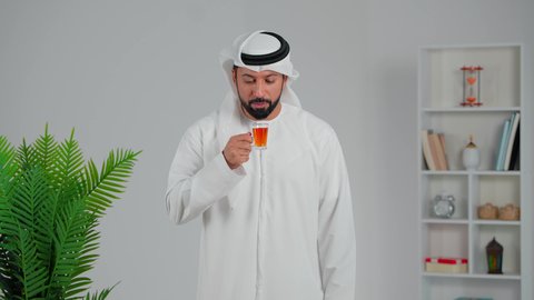 الاهتمام بالتدفئة الجسم بشكل جيد، محاولة الحصول على القليل من الراحة، اشارة الاعجاب والقبول، الاسترخاء وتناول مشروبات دافئة، بورتريه لرجل عربي خليجي اماراتي يرتدي الكندورة والغترة يقف ويمسك بيده كأس من الشاي، شاب  يقوم بالتلذذ بكوب من الشاي الساخن، خلفية رمادي