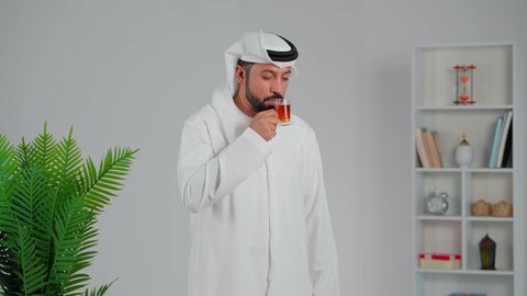 محاولة الحصول على القليل من الراحة، اشارة الاعجاب والقبول، الاسترخاء وتناول مشروبات دافئة، بورتريه لرجل عربي خليجي اماراتي يرتدي الكندورة والغترة يقف ويمسك بيده كأس من الشاي، شاب  يقوم بالتلذذ بكوب من الشاي الساخن، الاهتمام بالتدفئة الجسم بشكل جيد، خلفية رمادي