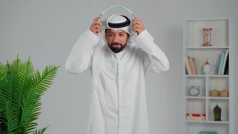 انجاز المهام عن بعد, التواصل مع الأهل والأصدقاء، استخدام تقنية الهيدفون، بورتريه مقرب لرجل عربي خليجي اماراتي يرتدي الكندورة والغترة يضع سماعه الرأس ينظر الى الكاميرا بإيماءات السرور، إجراء مكالمات صوتية، التطور التقني والتكنولوجي, خلفية رمادية