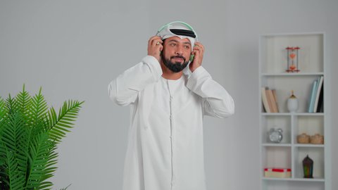 الاستمتاع بسماعة الموسيقى المفضلة, التواصل مع الأهل والأصدقاء، استخدام تقنية الهيدفون، بورتريه مقرب لرجل عربي خليجي اماراتي يرتدي الكندورة والغترة يضع سماعه الرأس ينظر الى الكاميرا بإيماءات السرور، إجراء مكالمات صوتية، التطور التقني والتكنولوجي, خلفية رمادية