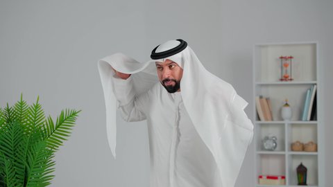 الوقوف باسقامة بايماءات السعادة والسرور, الاهتمام بالمظهر الخارجي, تصوير مقرب لرجل عربي خليجي اماراتي مبتسم يرتدي الكندورة و الغترة يعدل الغترة على رأسه، مفهوم الأناقة و الاهتمام بالجمال, مفهوم الرجولة والقوة والثقة بالنفس، خلفية رمادي