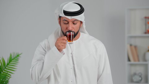 الاسترخاء وتناول مشروبات دافئة، محاولة الحصول على القليل من الراحة، بورتريه لرجل عربي خليجي اماراتي يرتدي الكندورة والغترة يقف ويمسك بيده كأس من الشاي، شاب  يقوم بالتلذذ بكوب من الشاي الساخن، الاهتمام بالتدفئة الجسم بشكل جيد، خلفية رمادي