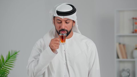 اشارة الاعجاب والقبول، الاسترخاء وتناول مشروبات دافئة، محاولة الحصول على القليل من الراحة، بورتريه لرجل عربي خليجي اماراتي يرتدي الكندورة والغترة يقف ويمسك بيده كأس من الشاي، شاب  يقوم بالتلذذ بكوب من الشاي الساخن، الاهتمام بالتدفئة الجسم بشكل جيد، خلفية رمادي