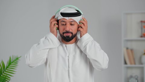 التواصل مع الأهل والأصدقاء، استخدام تقنية الهيدفون، انجاز المهام عن بعد, بورتريه مقرب لرجل عربي خليجي اماراتي يرتدي الكندورة والغترة يضع سماعه الرأس ينظر الى الكاميرا بإيماءات السرور، إجراء مكالمات صوتية، التطور التقني والتكنولوجي, خلفية رمادية