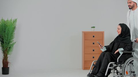 Taking commemorative selfies using a mobile phone, facilitating the movement of patients with motor disabilities, improving the psychological state of people with disabilities, sincerity and compassion between spouses, an Arab Gulf Emirati woman with special needs wearing an abaya and hijab sitting in a wheelchair with her husband