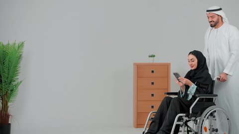 تصفح مواقع التواصل الالكتروني عبر الهاتف المحمول، تسهيل حركة المرضى ذوي الإعاقة الحركية، التحسين من نفسية أصحاب الهمم، الإخلاص والعطف بين الأزواج ، امراة عربيه خليجيه اماراتية من ذوى الاحتياجات الخاصة ترتدي العباءة و الحجاب تجلس على كرسي متحرك برفقه زوجها