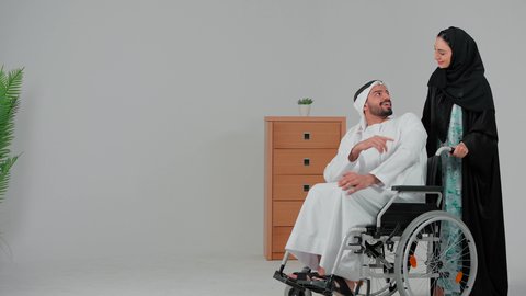 الوفاء والحنان المتبادل بين الزوجين، العنايه بأصحاب الهمم وتأمين احتياجاتهم ، نمط حياة ذوي الإعاقة الحركية ، تأمين أدوات ومعدات لمساعدة المرضي ، رجل خليجي من ذوي الاحتياجات الخاصه يجلس على كرسي متحرك ينظر الى زوجته، امراة عربيه خليجيه اماراتية تساعد زوجها على التنقل والحركة