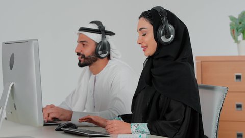 Emirati office jobs and professions, integrating work with modern and advanced technology, responding to inquiries and information, using headphone technology, a side view of an Emirati Gulf Arab woman wearing an abaya and hijab sitting in front of a computer working in the field of customer service and technical support with her colleague