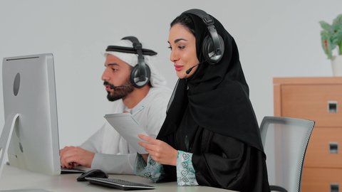Integrating work with modern and advanced technology, writing and taking notes, responding to and answering inquiries and questions, using headphone technology, a side view of an Emirati Gulf Arab woman wearing an abaya and hijab sitting in front of a computer working in the field of customer service and technical support with her colleague, Emirati office jobs and professions