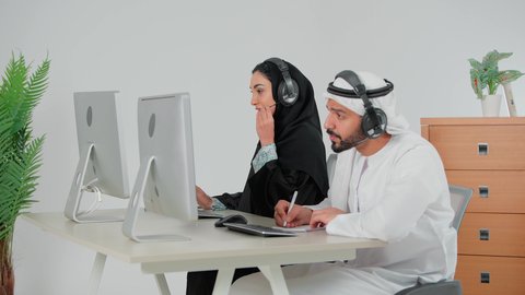 كتابة و تدوين الملاحظات، الرد والاجابة على الاستفسارات والاستعلامات، استخدام تقنية الهيدفون,  تصوير من الجانب لرجل عربي خليجي اماراتي يرتدي الكندورة و الغتره يجلس امام الحاسب الالى يعمل في مجال خدمة العملاء والدعم الفني برفقة زميلته, مهن ووظائف مكتبية اماراتية, دمج العمل مع التكنولوجيا الحديثة والمتطورة