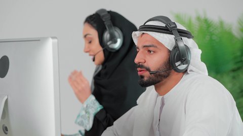 الرد والاجابة على الاستفسارات والاستعلامات، استخدام تقنية الهيدفون,  تصوير مقرب لرجل عربي خليجي اماراتي يرتدي الكندورة و الغتره يجلس امام الحاسب الالى يعمل في مجال خدمة العملاء والدعم الفني برفقة زميلته, مهن ووظائف مكتبية اماراتية, دمج العمل مع التكنولوجيا الحديثة والمتطورة