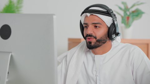 Responding to customer inquiries, using headphone technology, a close-up of an Emirati Gulf Arab man wearing a kandura and ghutra sitting in front of a computer working in customer service and technical support, Emirati office jobs and professions, integrating work with modern and advanced technology