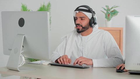 Using headphone technology, answering customer inquiries, a close-up of an Emirati Gulf Arab man wearing a kandura and ghutra sitting in front of a computer working in customer service and technical support, Emirati office jobs and professions, integrating work with modern and advanced technology