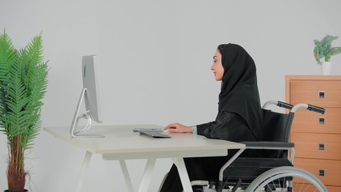 بيئة العمل المريحة في الشركات السعودية، تصوير من الجانب لامراه عربية خليجيه محجبه تجلس على كرسي متحرك تقوم  بمراجعة خطة العمل، رجل عربي خليجي اماراتي يرتدي الكندورة و الغتره يمسك بيده ملف ازرق، استخدام الحاسب والتقنية في مجال الاعمال